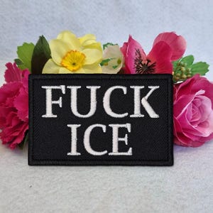 Pode incluir: Um patch retangular preto com as palavras "FUCK ICE" bordadas em branco. O patch é rodeado por um arranjo floral colorido, incluindo rosas cor-de-rosa, narcisos amarelos e folhas verdes.