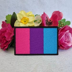 以下が含まれることがあります： 黒の縁取りのあるピンク、パープル、ブルーの刺繍パッチ。パッチはバイセクシュアルプライドフラッグを表しています。