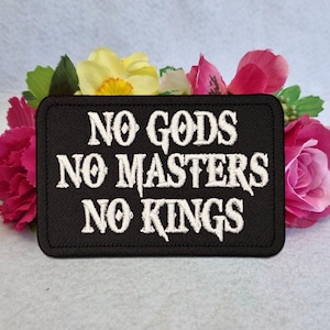 Könnte beinhalten: Ein rechteckiger, schwarz bestickter Aufnäher mit dem weißen Text NO GODS NO MASTERS NO KINGS in einer stilisierten Schriftart. Der Aufnäher hat einen gestickten Rand und liegt vor einem Bündel künstlicher rosa und gelber Blumen.