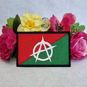 May include: Embroidered patch with a red and green diagonal design and a white anarchy symbol. The rectangular patch has a black border. The patch is surrounded by pink, yellow, and green flowers.