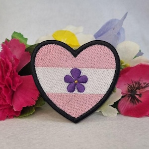 Puede incluir: Parche bordado en forma de corazón con un diseño a rayas rosas y blancas, una flor morada en el centro y un borde negro. El parche está rodeado de flores rosas y blancas.