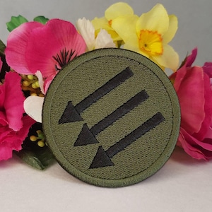 Puede incluir: Un parche bordado circular de color verde oliva que presenta tres flechas negras apuntando en diagonal hacia la izquierda. El parche tiene un borde cosido y está colocado frente a un grupo de flores artificiales de colores rosa y amarillo.