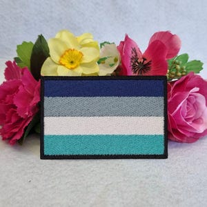 Puede incluir: Un parche bordado azul, gris, blanco y turquesa con un borde negro. El parche es una representación de la bandera del orgullo genderqueer.