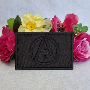 Op de afbeelding: Een zwart rechthoekig embleem met een gestikt ontwerp van een cirkel met de letters "A", "L" en "F" erin. Het embleem is omgeven door roze, gele en groene bloemen.