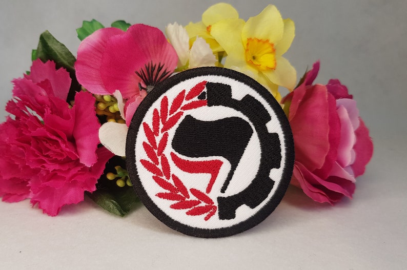May include: A black and white embroidered patch with a red stylized fist in a gear and a red laurel wreath. The patch is circular and has a white background.