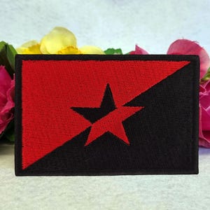 Op de afbeelding: Een rechthoekige patch met een rood en zwart sterontwerp. De patch is diagonaal verdeeld, met de linkerbovenhoek rood en de rechteronderhoek zwart. Een zwarte ster is gecentreerd op het rode gedeelte. Roze en gele bloemen op de achtergrond.