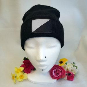 May include: Black beanie hat with a rectangular patch featuring a white and black diagonal design. The hat is displayed on a white mannequin head, adorned with colorful flowers. The hat is made of a soft, knitted material.