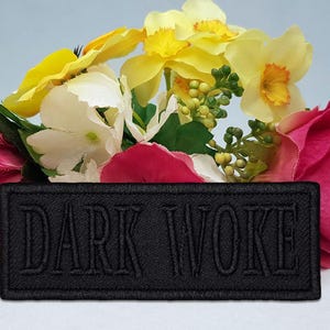 Op de afbeelding: Een zwart rechthoekig embleem met de geborduurde tekst "DARK WOKE" wordt getoond voor een kleurrijk bloemstuk. De bloemen omvatten roze, gele en witte bloemen.