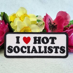 May include: A rectangular embroidered patch with a white background and a black border. The text reads I (with a red heart symbol) HOT SOCIALISTS in bold black letters. The patch is positioned in front of a cluster of yellow and pink flowers.