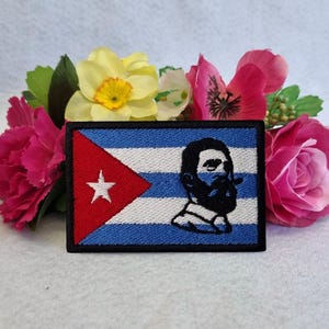 May include: Embroidered patch featuring the Cuban flag design with a red triangle, white star, blue stripes, and a portrait of a man with a beard. The patch is surrounded by pink and yellow flowers.