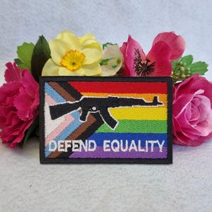 Puede incluir: Un parche rectangular bordado que presenta la silueta de un rifle negro centrada sobre un fondo de bandera progress pride con franjas horizontales de color rojo, naranja, amarillo, verde, azul y violeta, además de blanco, rosa, azul claro, marrón y negro. El texto DEFEND EQUALITY está bordado en blanco en la parte inferior.
