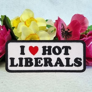 May include: A rectangular embroidered patch with a white background and a black border. The text reads I (with a red heart symbol) HOT LIBERALS in bold black lettering. The patch is positioned in front of a cluster of yellow and pink flowers.