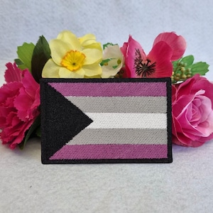 May include: A black, white, grey, and purple embroidered patch with a black triangle pointing to the left. The patch is surrounded by pink, white, and yellow flowers.