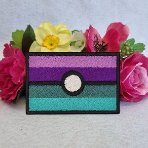 Könnte beinhalten: Gestickter Aufnäher mit einem Design aus horizontalen Streifen in Lila, Türkis und Grün, mit einem weißen Kreis in der Mitte. Der Aufnäher ist schwarz umrandet und von rosa und gelben Blumen umgeben.