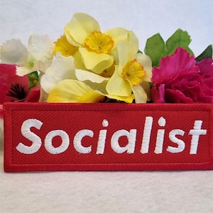 May include: A rectangular red embroidered patch featuring the word Socialist in white capital letters. The patch is positioned in front of a cluster of artificial flowers, including yellow daffodils, white blossoms, and vibrant pink carnations.