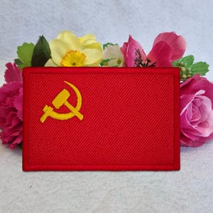 May include: A rectangular red embroidered patch featuring a yellow hammer and sickle symbol in the upper left corner. The patch has a visible diagonal fabric weave texture and a stitched border, presented against a background of colorful flowers.