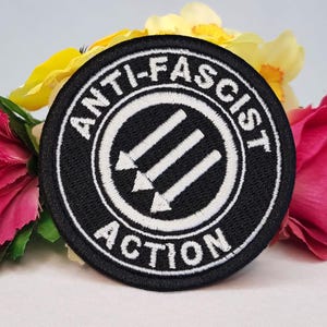 May include: A circular embroidered patch featuring a black background with white stitching. The design includes three downward-pointing arrows inside a circle, surrounded by the text ANTI-FASCIST ACTION in a bold, white, capitalized sans-serif font.