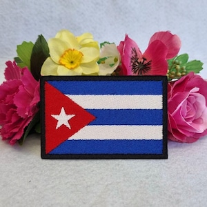 May include: A rectangular patch featuring the Cuban flag, with a red triangle and white star on the left, and blue and white stripes on the right. The patch is surrounded by a black border and set against a backdrop of pink, yellow, and green flowers.