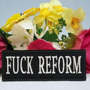 Peut inclure: Un écusson rectangulaire noir avec les mots "FUCK REFORM" en lettres blanches brodées. L'écusson est placé sur un fond de fleurs colorées, notamment des fleurs roses, jaunes et blanches.