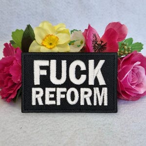 Op de afbeelding: Een zwart embleem met witte geborduurde tekst "FUCK REFORM" wordt omringd door kleurrijke bloemen. De bloemen omvatten roze rozen, gele narcissen en andere bloesems.