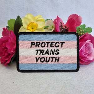 Może przedstawiać: Naszywka haftowana w kolorze niebieskim, białym i różowym z tekstem "PROTECT TRANS YOUTH" na czarnym tle.
