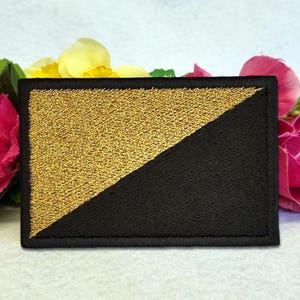 Puede incluir: Un parche rectangular bordado que presenta un diseño dividido en diagonal. El triángulo superior izquierdo está relleno con hilo dorado metálico, mientras que el triángulo inferior derecho está relleno con hilo negro sólido. Tiene un borde negro cosido.