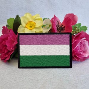 May include: A pink, white, and green embroidered patch with a black border. The patch is surrounded by pink, white, and yellow flowers.