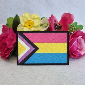 May include: A pink, yellow, and blue embroidered patch with a white and purple chevron design. The patch is surrounded by pink and yellow flowers.