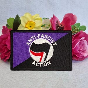 May include: A rectangular embroidered patch featuring a diagonal split design with purple on the top left and black on the bottom right. A central white circle contains a black and red flag icon with the text ANTI-FASCIST ACTION in white.