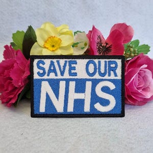 May include: A blue and white embroidered patch with the text "Save Our NHS" surrounded by pink and yellow flowers.
