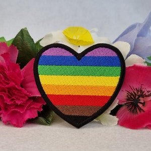 May include: A rainbow pride heart-shaped patch with a black outline. The patch is made of embroidered fabric and has six stripes of color: purple, blue, green, yellow, orange, and red. The bottom stripe is brown.