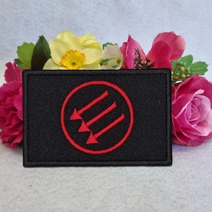 Pode incluir: Um patch retangular preto com um círculo vermelho contendo três setas apontando para baixo. O patch é cercado por flores rosa, amarelas e verdes, sugerindo um símbolo ou emblema.