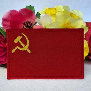 Könnte beinhalten: Roter gestickter Aufnäher mit einem goldenen Hammer-und-Sichel-Symbol.
