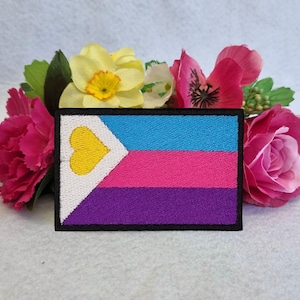 Könnte beinhalten: Ein gestickter Aufnäher mit einer mehrfarbigen Flagge mit einem gelben Herzen in der oberen linken Ecke. Die Flagge hat blaue, rosa und violette Streifen.