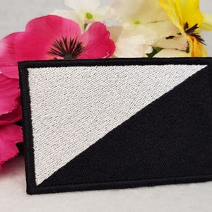 May include: A black and white embroidered patch with a diagonal line dividing the patch. The patch is surrounded by a black border.