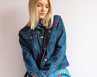 May include: A blue denim jacket with a brown leather strap and a blue denim skirt with embroidered flowers in red, yellow, and white.