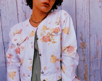 May include: A white denim jacket with a floral print. The jacket has a button closure and a collar. The model is wearing a green and white plaid skirt.