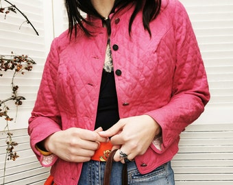 May include: A pink quilted jacket with a mandarin collar and button closure. The jacket is worn over a black top and blue jeans. A brown leather backpack is worn over the shoulder.