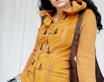 May include: A mustard yellow suede jacket with a knitted hood and long sleeves. The jacket has a zipper closure and decorative buttons. The jacket is worn over a black and white plaid skirt.