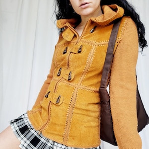 Könnte beinhalten: Eine senfgelbe Wildlederjacke mit Strickkapuze und langen Ärmeln. Die Jacke hat einen Reißverschluss und dekorative Knöpfe. Die Jacke wird über einem schwarz-weiß karierten Rock getragen.