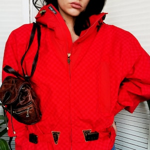 Puede incluir: Chaqueta roja con capucha, de patrón a cuadros y cierre de cremallera. La chaqueta tiene dos bolsillos con hebillas y la palabra "GORE-TEX" es visible en el hombro izquierdo.
