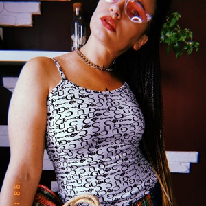Puede incluir: Una mujer con un top blanco de tirantes con un patrón blanco y negro de la palabra "Gucci" y un collar de cadena. También lleva pantalones de cuadros y tiene trenzas negras largas. Lleva un bolso de cesta tejido.