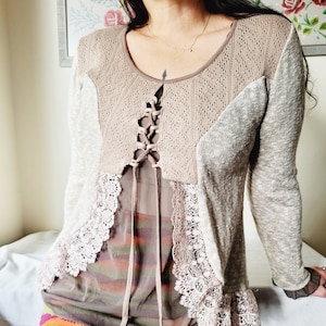 Peut inclure: Un pull en tricot beige avec une bordure en dentelle et un détail de laçage à l'avant. Le pull est superposé à un haut transparent à rayures brunes et orange.
