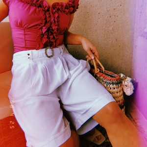 May include: A magenta ruffled top paired with white high-waisted shorts and a woven handbag. The top features a square neckline and a fitted silhouette. The shorts have a belted waist with a decorative buckle. The handbag has a colorful design.