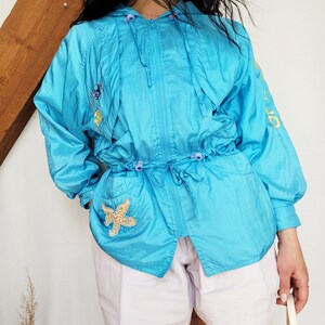 May include: A turquoise hooded jacket with a drawstring waist and zipper closure. The jacket features embroidered sea-themed patches, including a starfish. The sleeves have yellow text. The model is wearing white shorts.