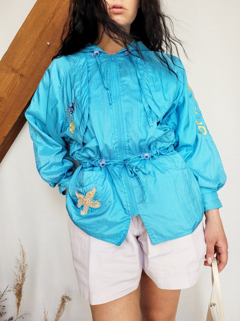 May include: A turquoise hooded jacket with a drawstring waist and zipper closure. The jacket features embroidered sea-themed patches, including a starfish. The sleeves have yellow text. The model is wearing white shorts.