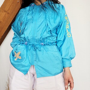 May include: A turquoise nylon jacket with a drawstring waist, hood, and zipper closure. The jacket features a starfish patch on the pocket and the words "A WORLD" embroidered in yellow on the sleeve. The jacket is paired with light purple shorts.