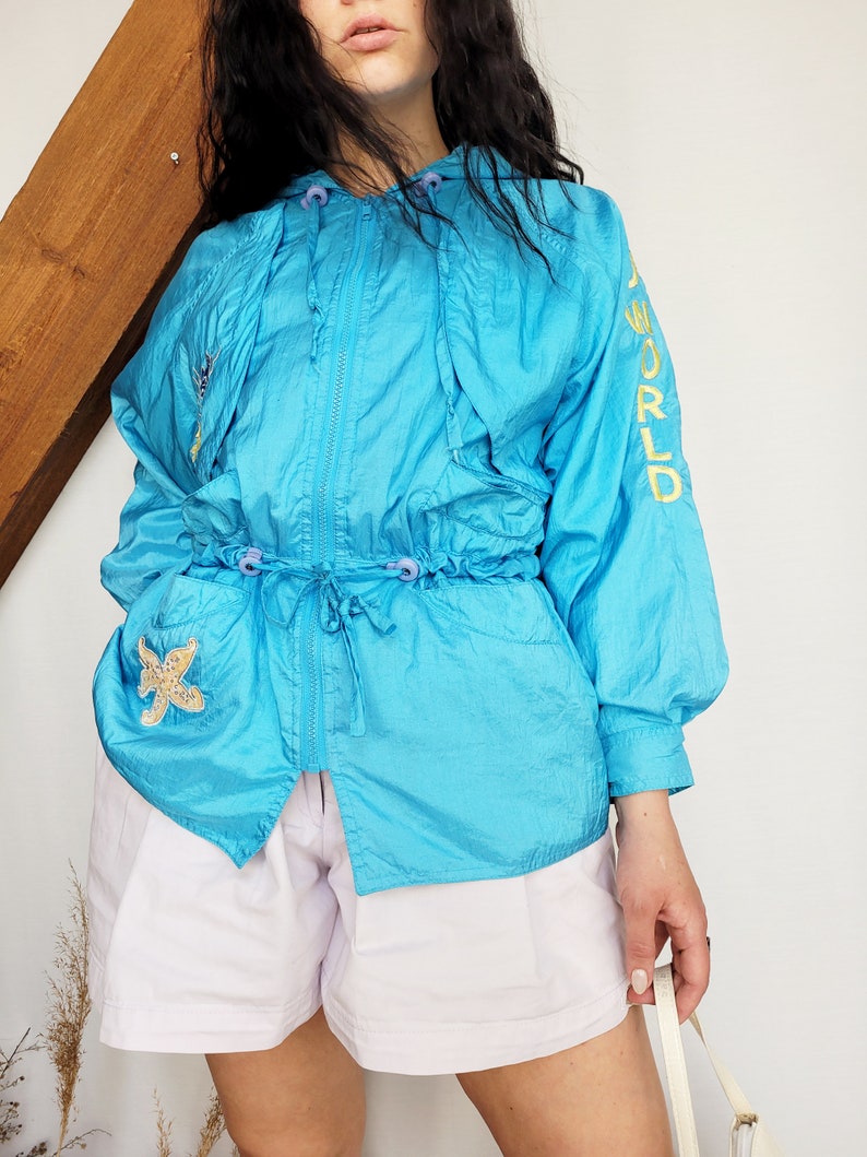 May include: A turquoise nylon jacket with a drawstring waist, hood, and zipper closure. The jacket features a starfish patch on the pocket and the words "A WORLD" embroidered in yellow on the sleeve. The jacket is paired with light purple shorts.
