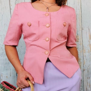 May include: A pink blazer with a round neckline and gold buttons. The blazer has two pockets on the front and is cropped at the waist.