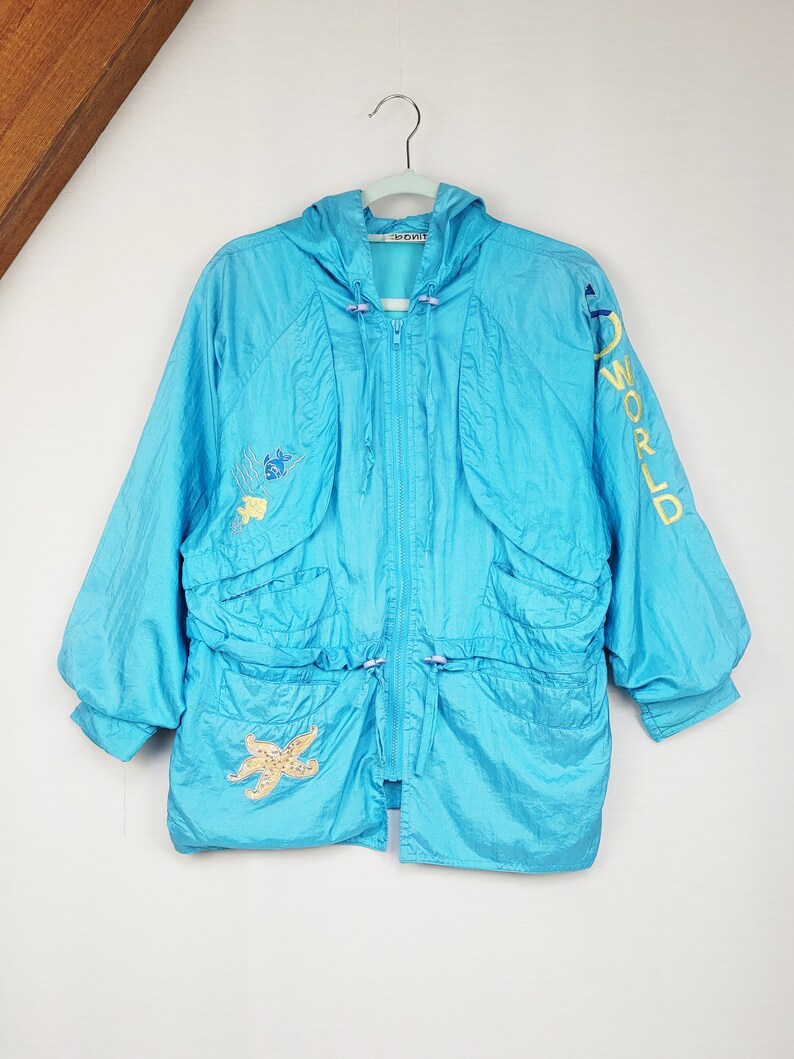 May include: A turquoise hooded jacket with a zipper closure, drawstring waist, and two front pockets. The jacket features embroidered starfish and aquatic designs, with "WORLD" in yellow lettering on the sleeve. The jacket is hanging on a white hanger.